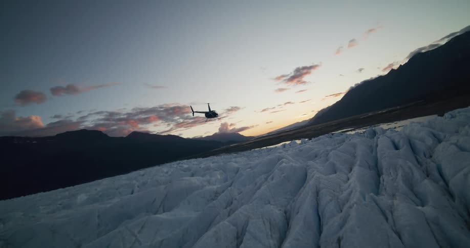 A cinematic slow-motion shot follows a helicopter flying over a vast glacier before coming in for a landing at sunset