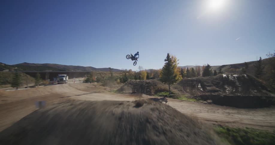 Dynamic slow-motion shot follows a motocross rider launching off a massive dirt jump against a clear blue sky