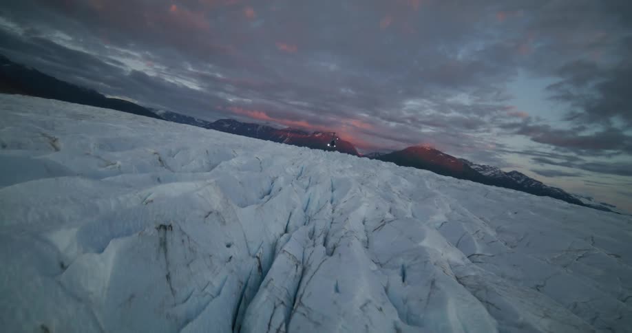 An aerial view pulls away from a helicopter landed on a vast Alaskan glacier at sunset, revealing the epic mountain landscape