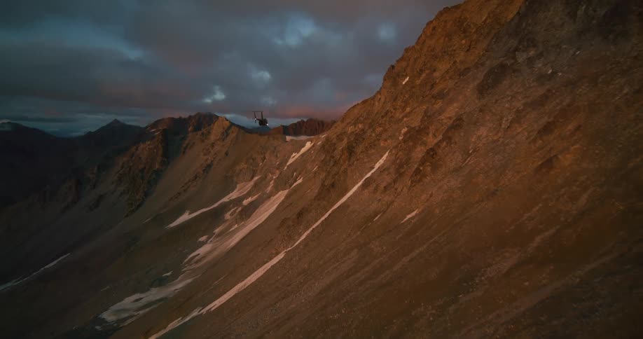 An aerial view of a helicopter flying alongside a rugged mountain range during a dramatic sunset