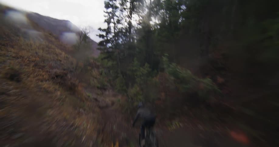 An exhilarating FPV drone shot follows a mountain biker speeding down a steep and winding dirt trail through a moody forest