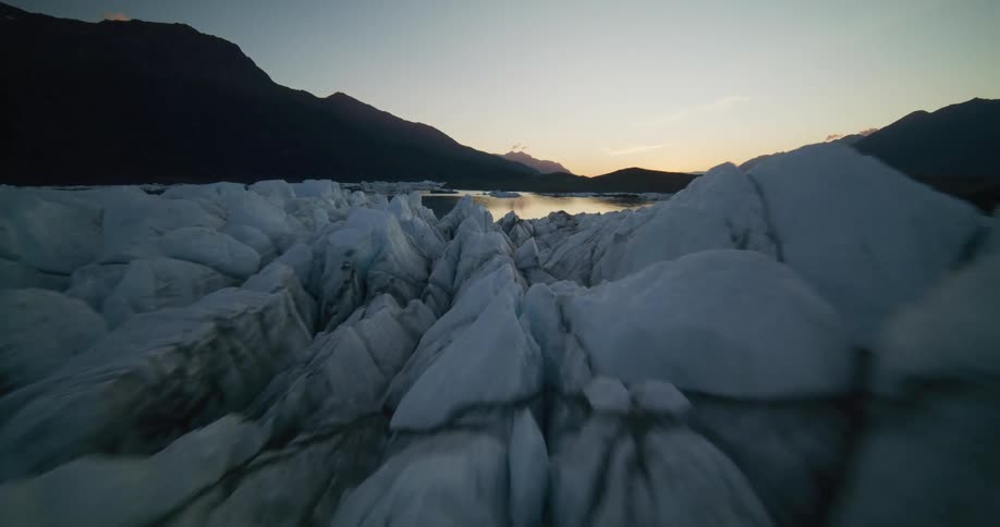 A slow aerial push-in over a vast, cracked glacier towards a calm lake surrounded by mountains at dusk