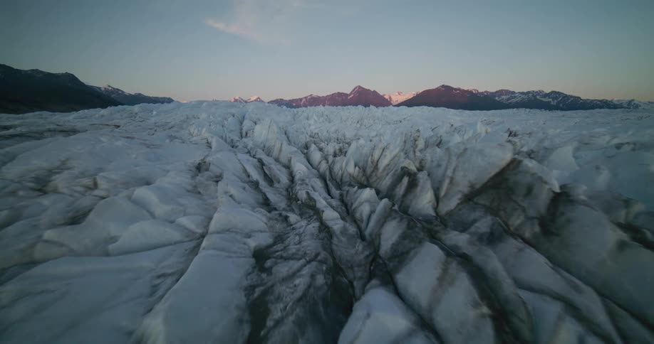 A stunning aerial flight from a tranquil lake over the vast, rugged surface of a massive glacier towards sun-kissed mountains at twilight