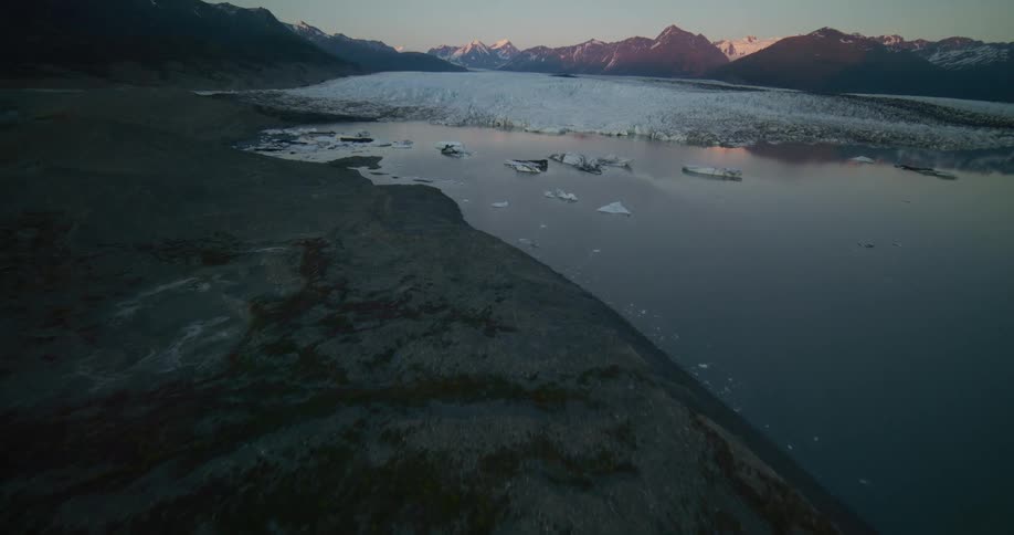 A serene aerial flight over a dark, rocky landscape reveals a massive glacier and floating icebergs in a calm lake at twilight