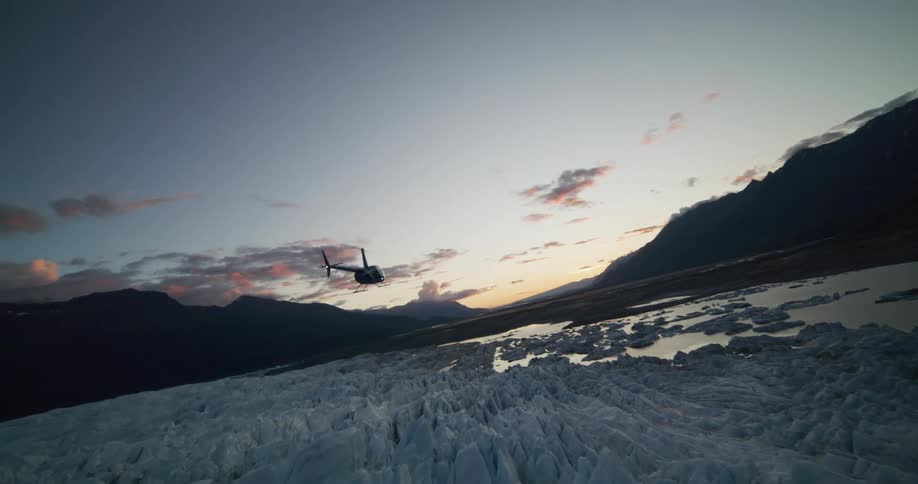 A helicopter flies low across a vast, frozen glacier against a stunning sunset sky and mountain backdrop