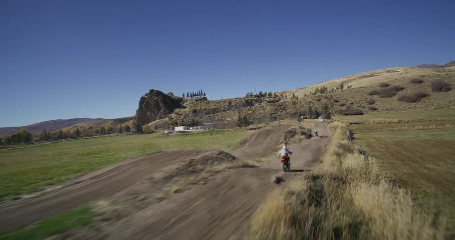 Drone follows several motocross riders as they race and jump along a dirt track in the countryside on a clear sunny day