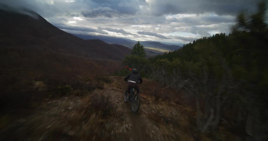 Dynamic follow-cam footage captures a mountain biker descending a steep, winding trail through a vast mountain valley under a dramatic, cloudy sky