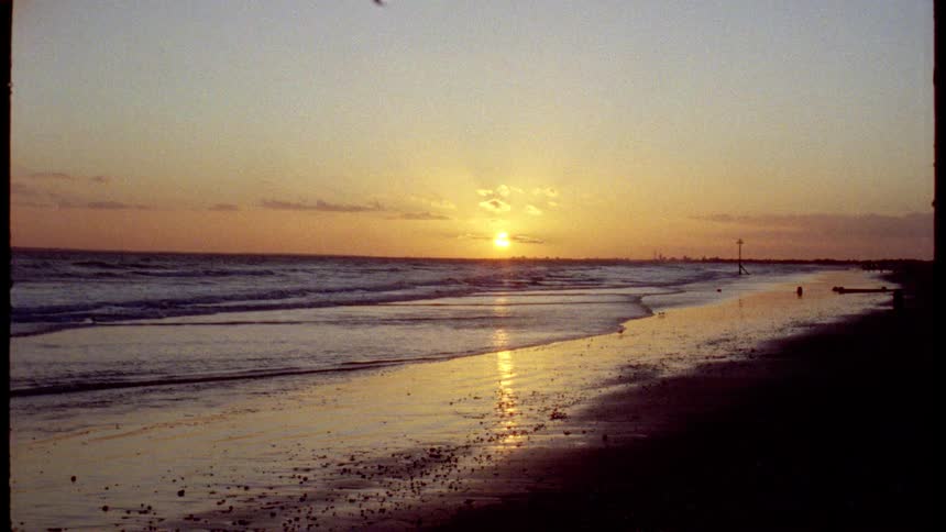A vintage film captures the golden sunset reflecting on the wet sand as waves gently crash on the shore