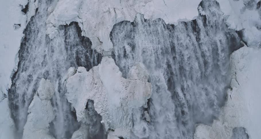 A majestic high-angle slow-motion view shows a powerful waterfall cascading through an icy, snow-covered landscape