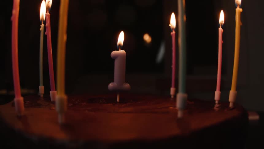 A number one candle and other colorful candles flicker on a chocolate cake celebrating a first birthday in a dark room