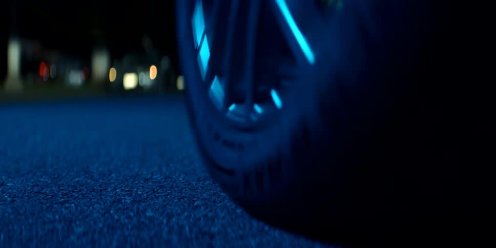 A cinematic low-angle shot captures a car's tire rolling into frame and parking on a blue-lit street at night