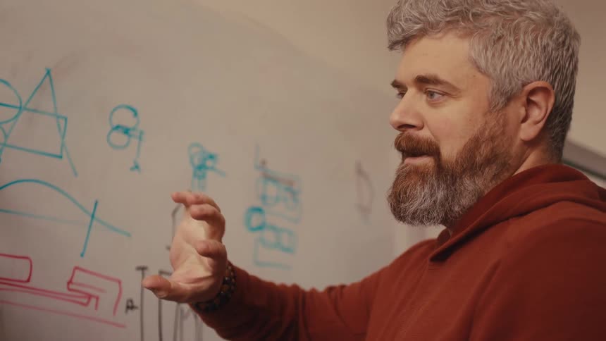 A bearded engineer in a hoodie explains a new product prototype while gesturing towards diagrams on a whiteboard