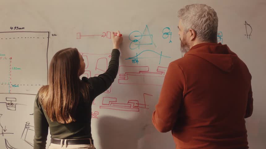 A man and a woman collaborate on a design, drawing intricate plans and diagrams on a whiteboard