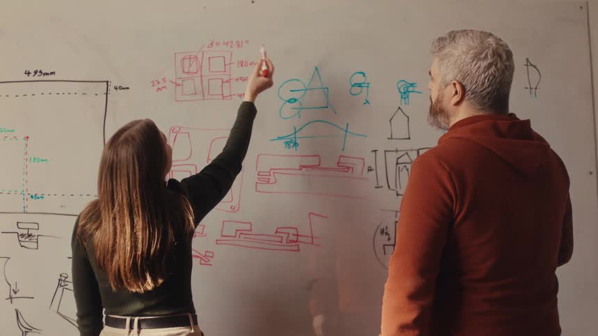 A young female engineer and her senior colleague brainstorm and sketch project schematics on a whiteboard in a design studio
