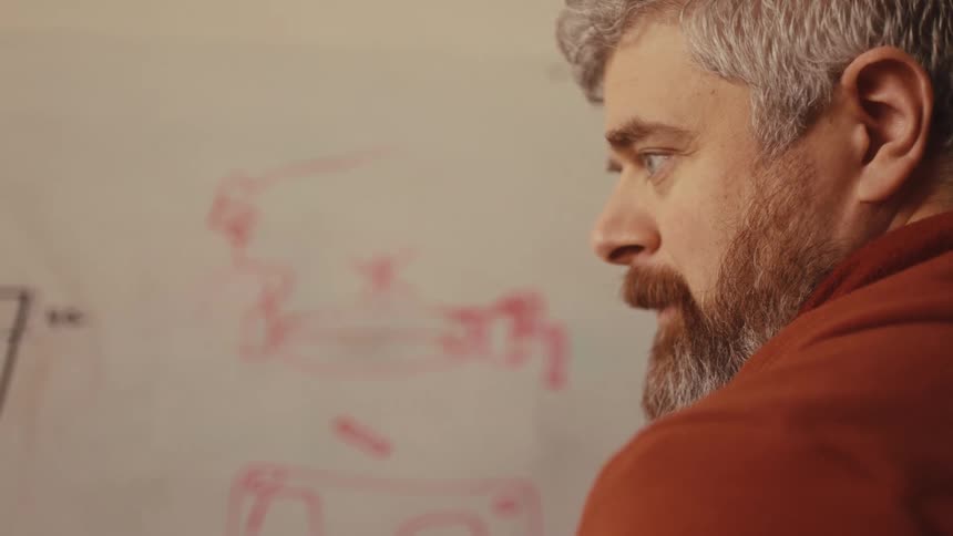 An engineer with a beard sketches a technical diagram on a whiteboard before assembling a prototype with his colleague
