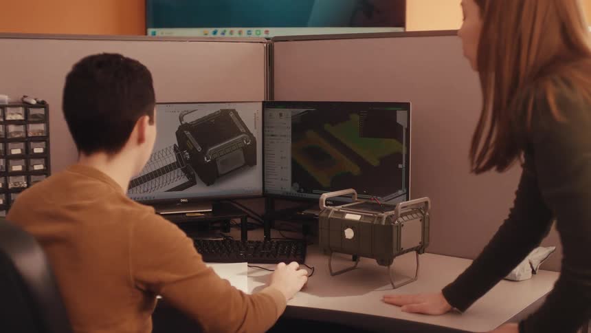 Two engineers collaborate on a product design, comparing the 3D model on the computer with a physical prototype on the desk