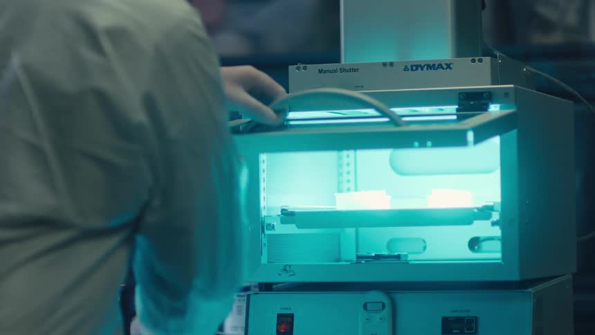 A laboratory technician in a protective suit carefully places and adjusts samples inside a glowing blue UV light curing chamber