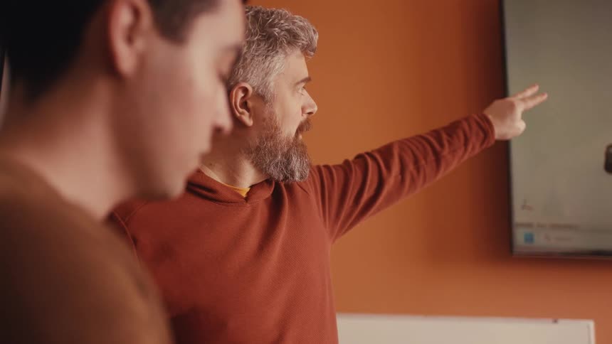 A senior male colleague with a beard points to a screen, passionately explaining a concept to his younger coworker in a modern office