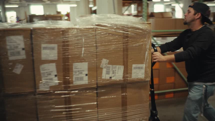 Tracking shot of a male warehouse worker pushing a pallet jack loaded with a large stack of boxes through the facility