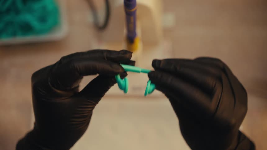 A worker wearing black gloves inspects a small, turquoise plastic part in a workshop