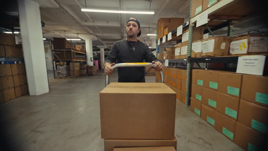 First-person perspective of a warehouse worker pushing a dolly loaded with cardboard boxes down an aisle