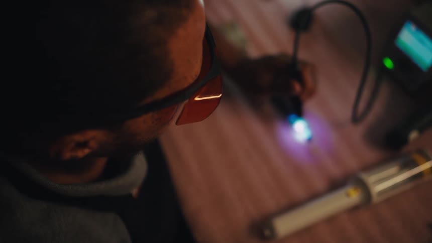 An engineer in protective glasses meticulously inspects a glowing electronic circuit board with a lighted tool on a workbench