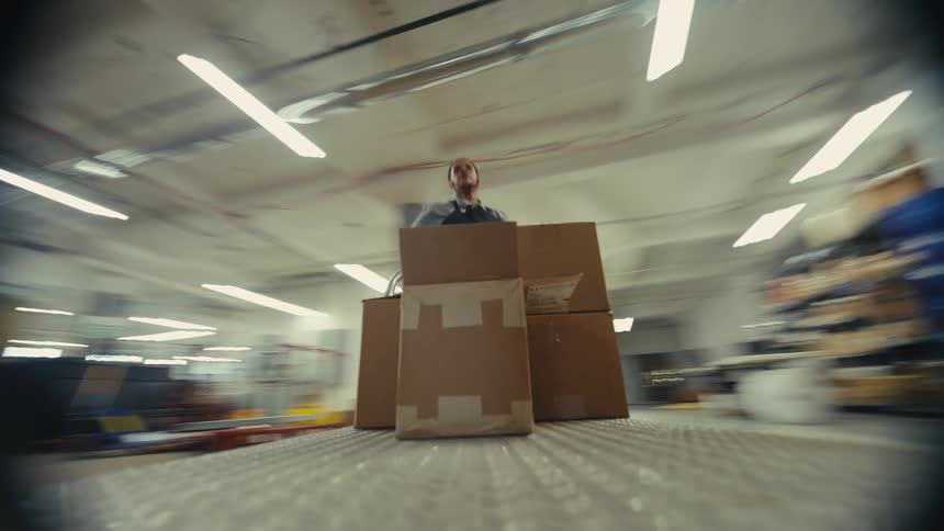 A low-angle time-lapse shot shows a worker pushing a cart loaded with boxes through a bustling factory warehouse