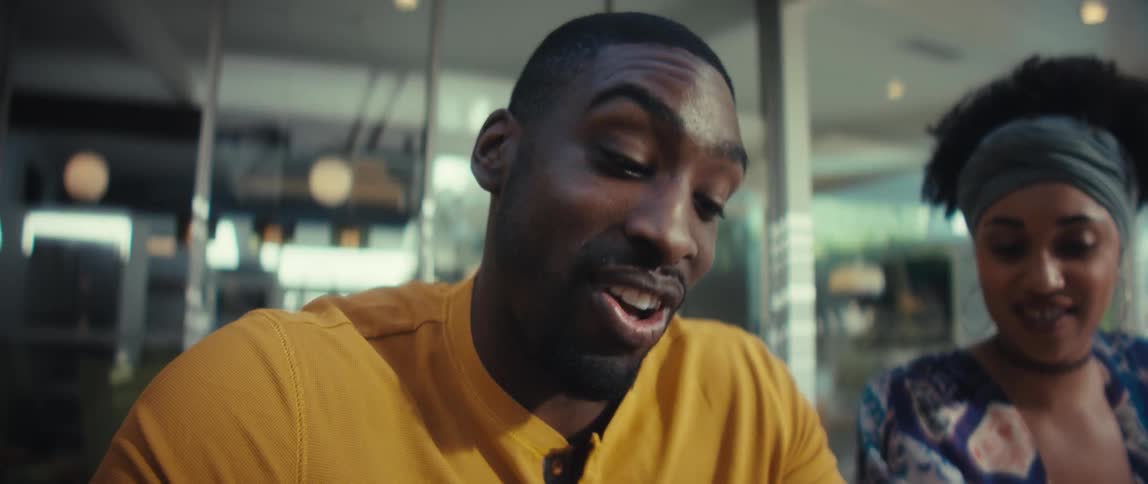 A Black man and woman joyfully collaborate on their finances, using a calculator and laptop at a wooden table in a sunlit room