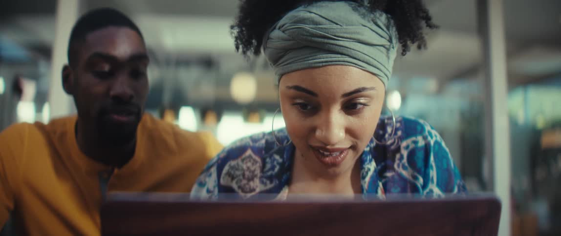A stylish young Black woman reacts with shock and then pure joy while looking at a laptop with her partner beside her
