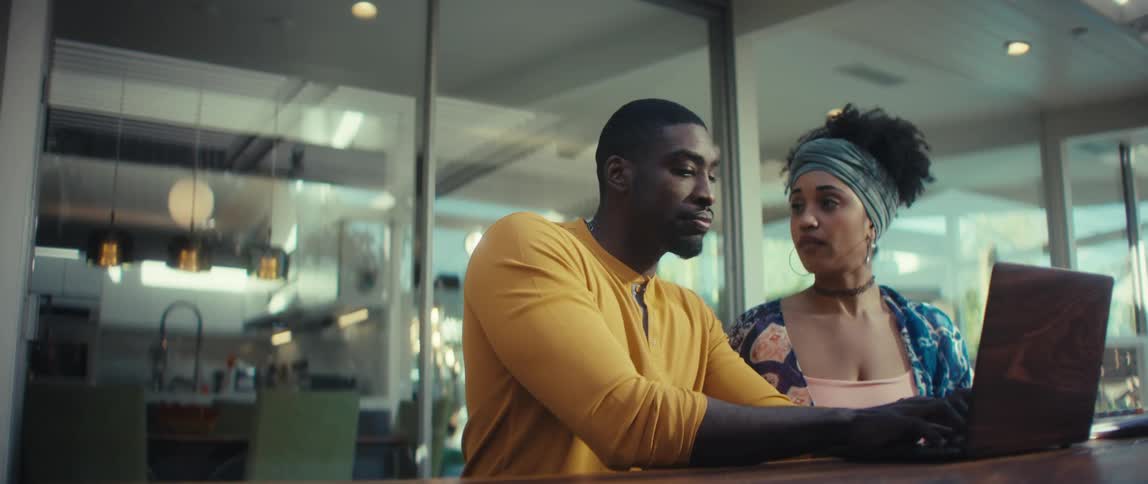 A young Black couple collaborates on a project, opening a laptop together and working in their modern sunlit home