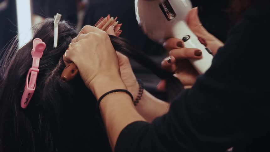 In slow motion, a professional hairstylist blow-dries a model's long dark hair with a round brush backstage