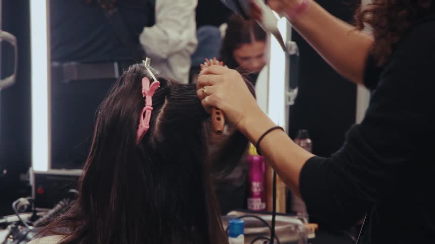 A professional hairstylist uses a round brush and hair dryer to style a woman's long dark hair in a salon