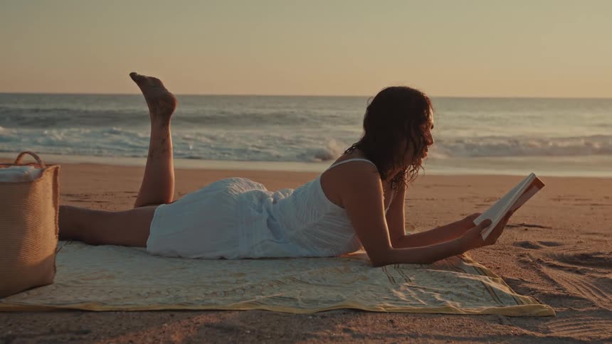 A woman in a white dress peacefully reads a book while lying on a blanket on the beach at sunset