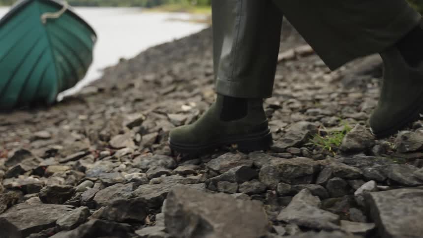 A person wearing fashionable green suede boots walks in slow motion over a rocky shoreline next to a lake and a small boat