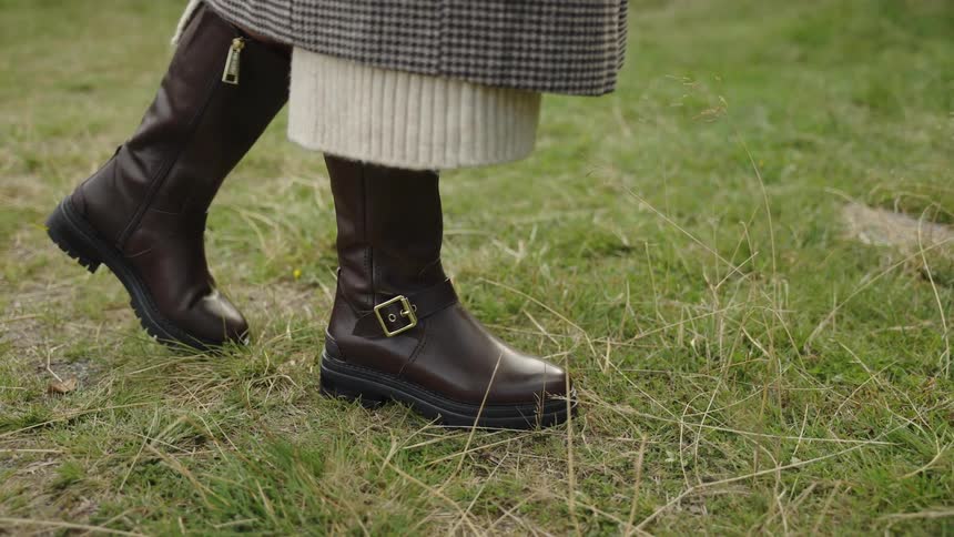 A close-up of a stylish woman's brown leather boots as she walks across a grassy field