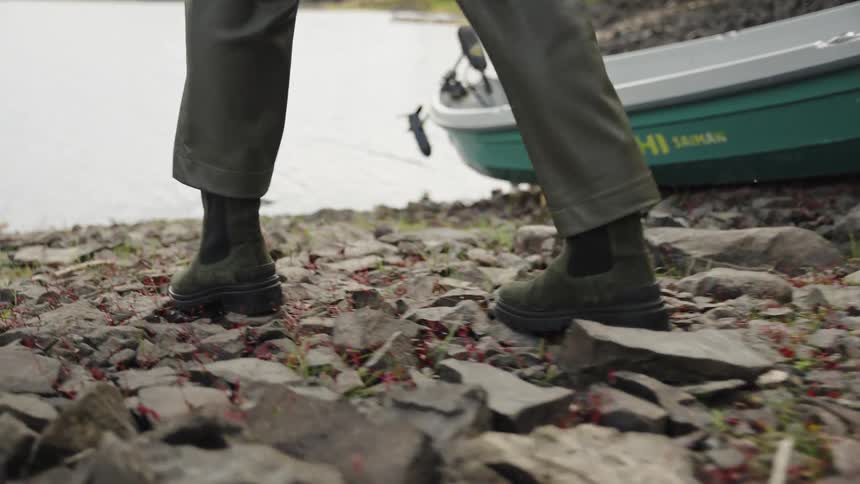A close-up slow-motion view of stylish green boots walking across a rocky shoreline by a lake