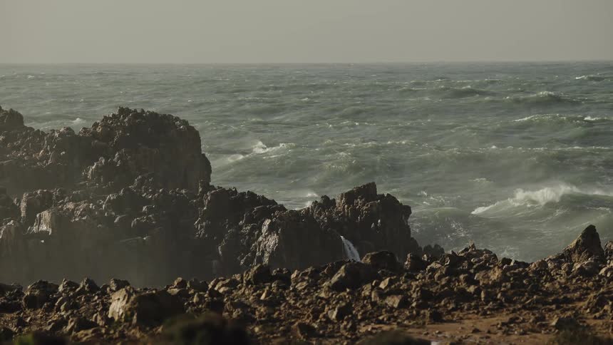 A slow-motion wide shot captures the turbulent ocean as powerful waves crash against a rugged, rocky shoreline