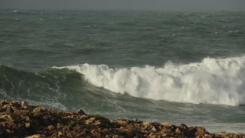 A powerful ocean wave builds and crashes in dramatic slow motion, viewed from a rocky coastline under an overcast sky