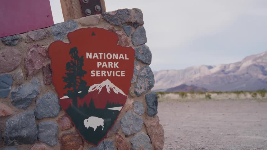A National Park Service sign on a rustic stone pillar stands before a vast desert and mountain landscape