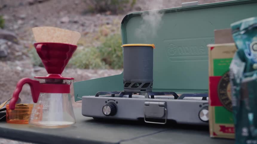 Water boils in a pot on a portable camp stove next to a pour-over coffee maker at an outdoor campsite