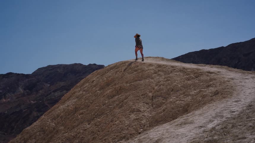 A lone hiker in a hat walks along a dusty ridge, surrounded by the arid, colorful mountains of a desert park