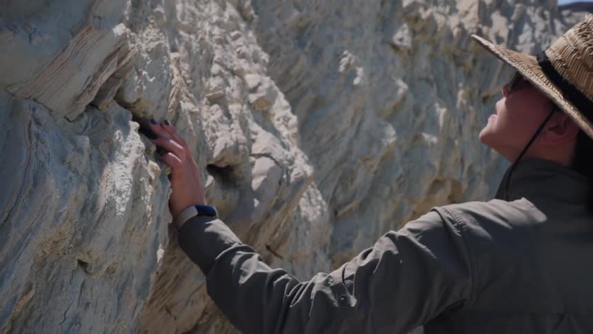 A curious female explorer in a sun hat gently touches the unique texture of a large rock formation in slow motion before walking away