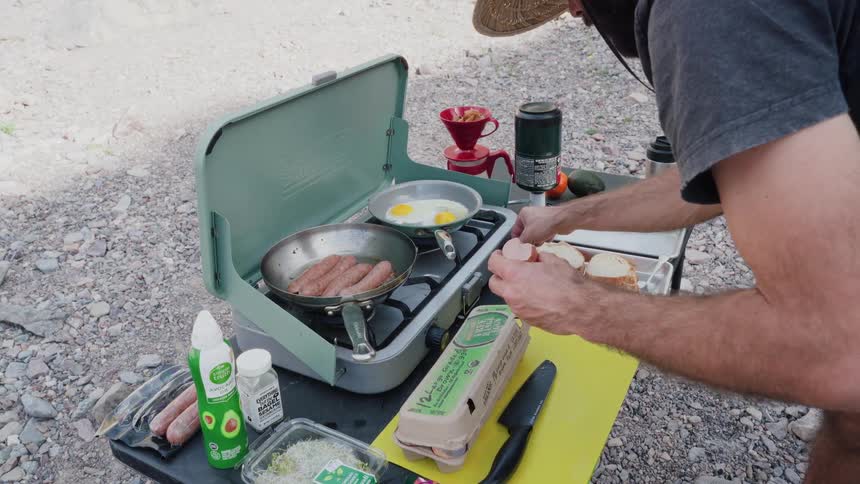 A man cracks eggs into a pan while cooking a sausage breakfast on a portable camp stove outdoors