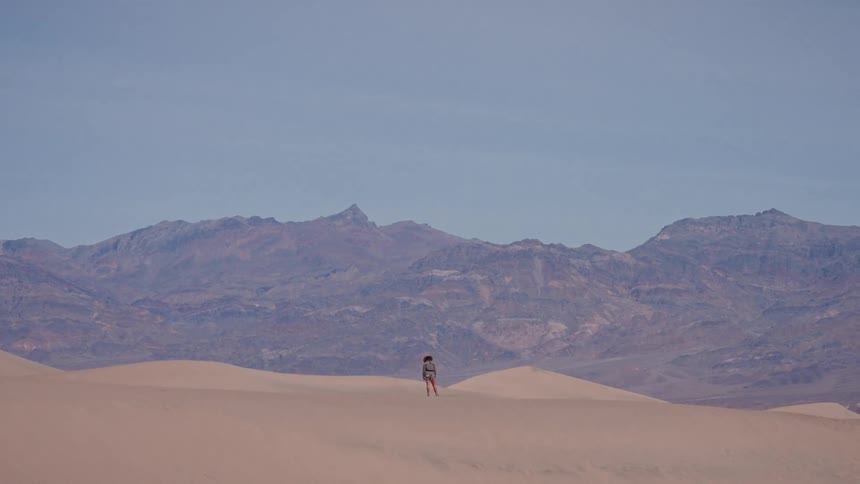 A lone adventurer stands still on a sweeping sand dune, dwarfed by the immense desert landscape and distant mountains