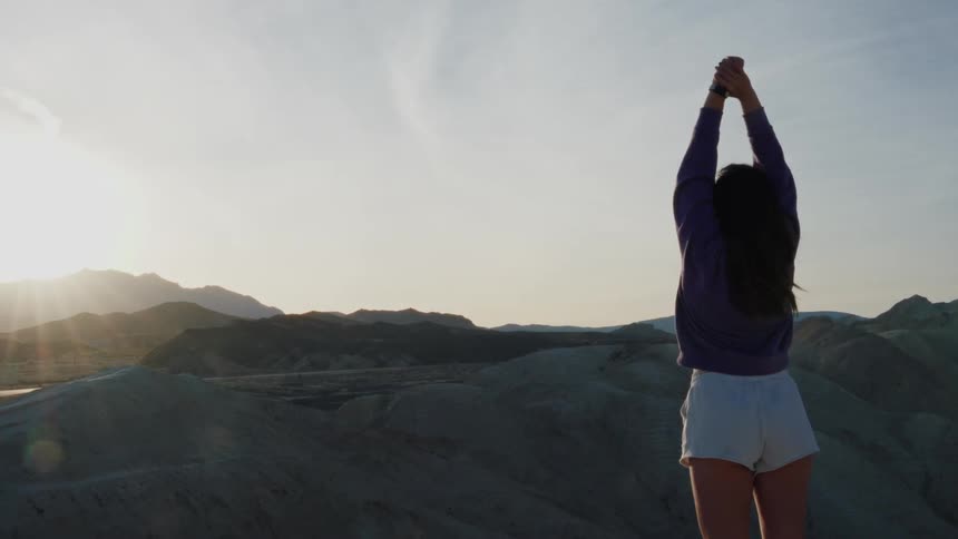 A young woman stands on a mountain peak, stretching and raising her arms to the sky as she overlooks a vast desert landscape at sunset