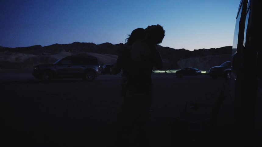 A silhouetted couple romantically embraces and kisses in the desert landscape during a road trip at twilight