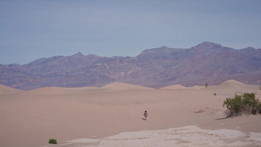 A lone traveler hikes across vast sand dunes with a mountain range in the distant background