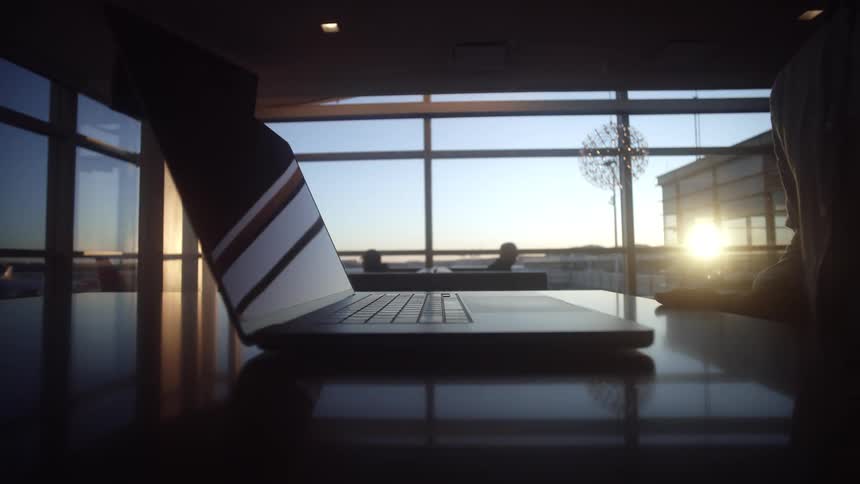 A person is silhouetted as they open and close their laptop on a reflective desk in an airport lounge at sunset