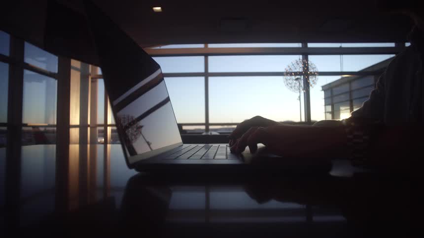 Silhouette of a person's hands typing on a laptop on a reflective desk in a modern lounge at sunset