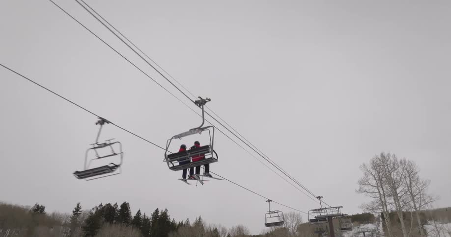 Two skiers ride a chairlift up a snowy mountain over a forest of bare trees on an overcast day